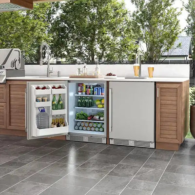 A built-in outdoor refrigerator by Zephyr, seamlessly integrated into a wood outdoor kitchen island. One of the compact stainless steel units is shown with its door open, revealing organized shelves filled with a variety of cold beverages and fresh food items, while the adjacent unit remains closed. The setup includes a counter-top grill, prep space, and a sink on a tiled patio.