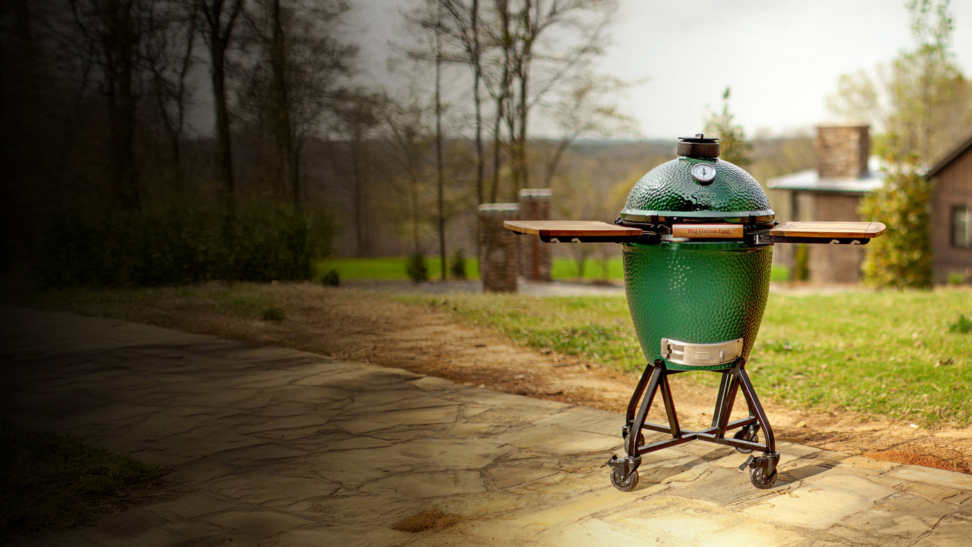 Big Green Egg ceramic charcoal grill with side shelves on a rolling stand, set on a stone patio with a grassy backdrop