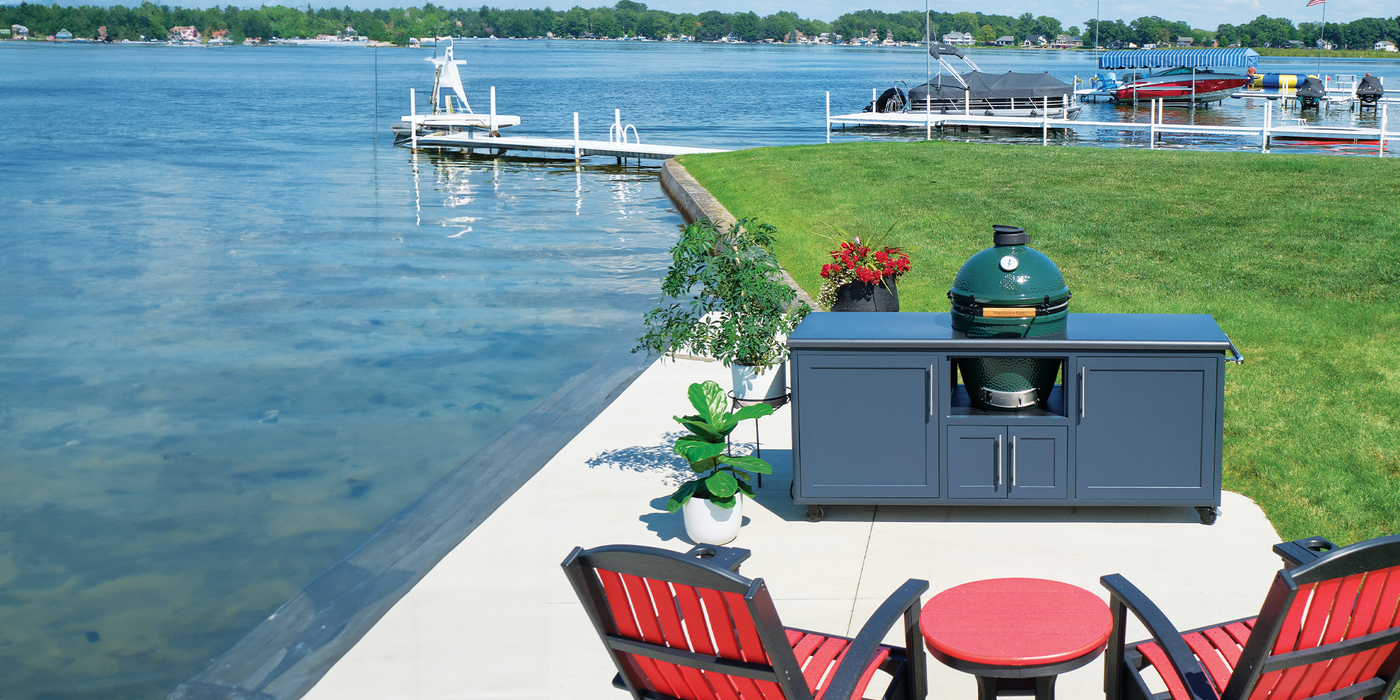 Big Green Egg ceramic grill installed in a Challenger Designs outdoor kitchen cabinet on a lakeside patio with red Adirondack chairs and dock access.