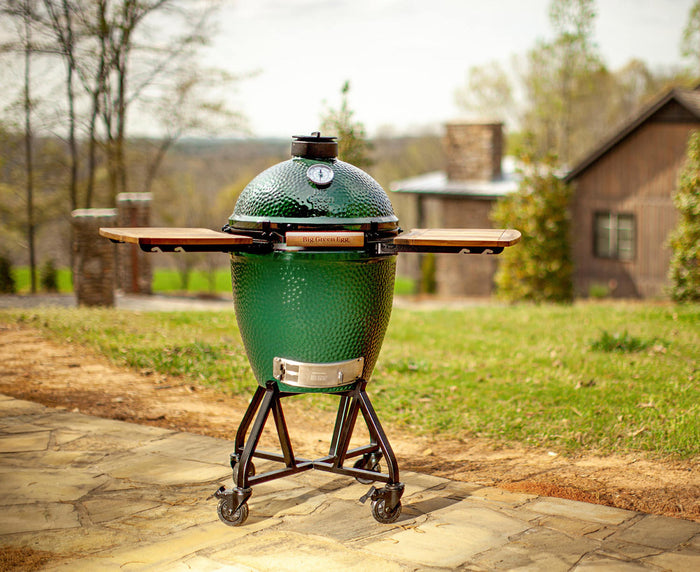 Big Green Egg ceramic charcoal grill with folding side shelves on a rolling cart, set outdoors on a stone patio.
