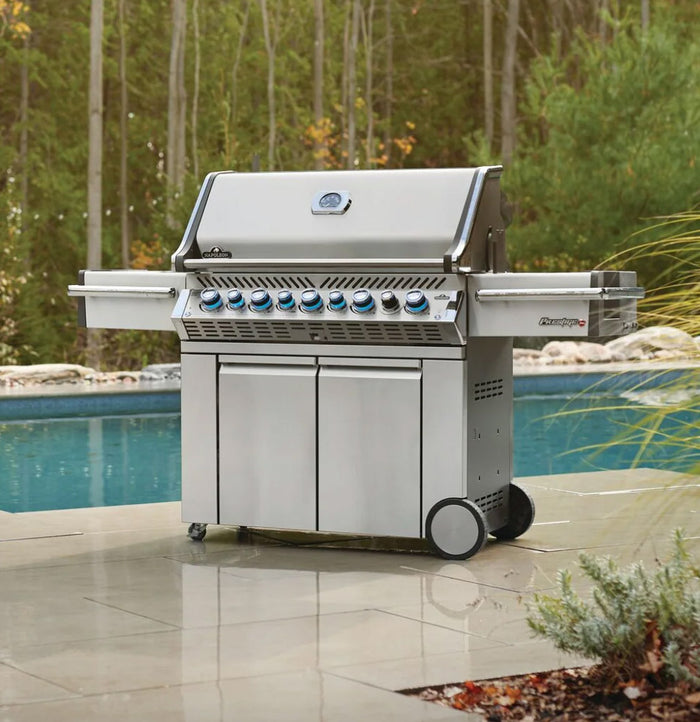 Napoleon Prestige Pro stainless steel gas grill with illuminated knobs and dual side shelves, set poolside on a patio.