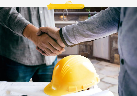 Two men shaking hands over outdoor kitchen plans with a yellow construction helmet in the foreground.