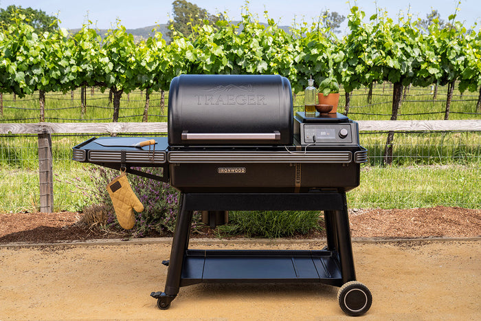 Traeger Ironwood pellet grill set up outdoors near a vineyard with side shelf, touchscreen controls, and grilling tools.