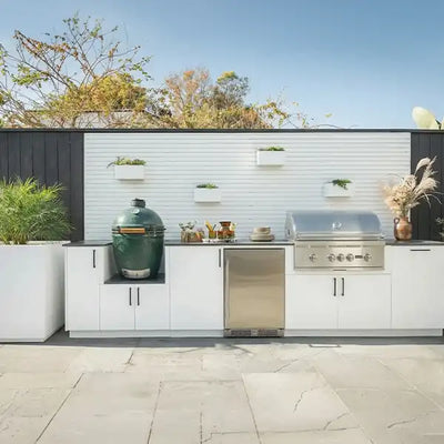 An Urban Bonfire custom modular outdoor kitchen featuring a white slat wall, dark countertops, a large integrated Big Green Egg kamado grill, a Coyote built-in gas grill, a stainless steel refrigerator, and ample cabinet storage. The modern setup includes white vertical planters and is situated on a stone patio.