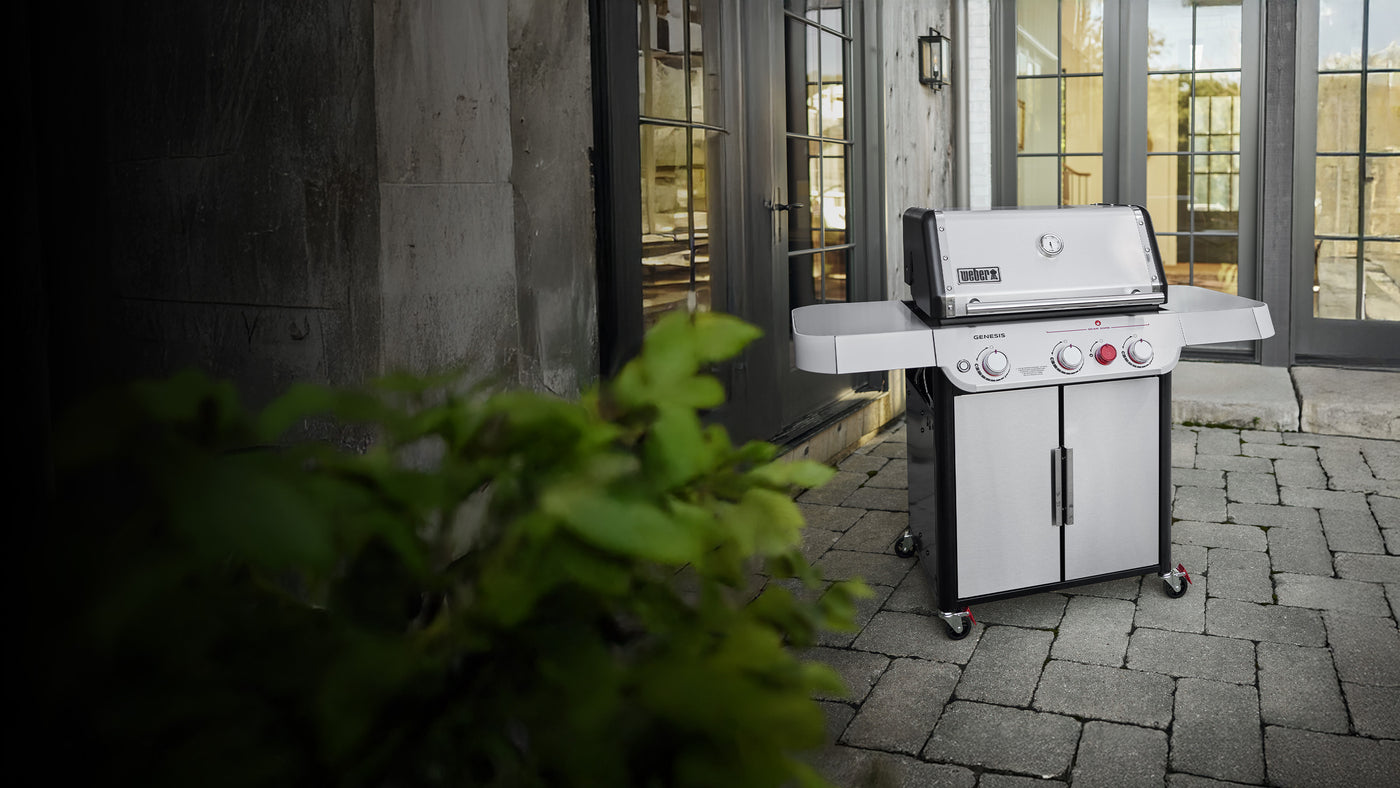 Weber Genesis SP-S-325 stainless steel gas grill on a stone patio, positioned in front of modern double glass doors.