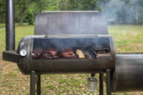 3 briskets being smoked on an aaron franklin barbecue pit offset smoker, with smoke and vapor surrounding the grill, in front of a grassy woods background, this grill is available now at texas star grill shop